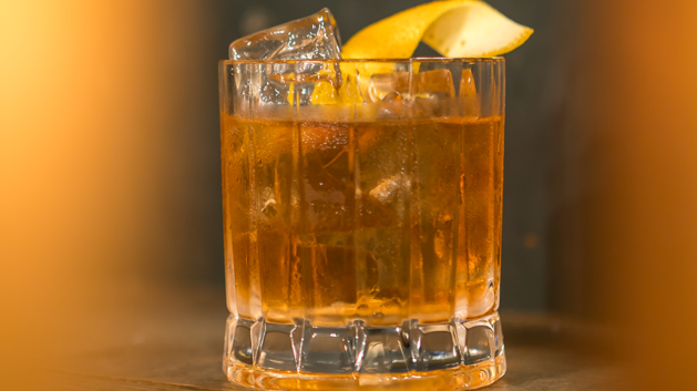 Imagem de um copo de vidro com o drink Rusty Nail, gelo e um twist de laranja sobre uma mesa de madeira, com efeitos de foto em laranja nas duas bordas laterais.