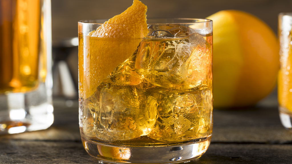 Imagem de um copo de vidro com o drink Rusty Nail, gelo e um twist de laranja sobre uma superfície escura, com uma laranja descascada pela metade ao fundo.